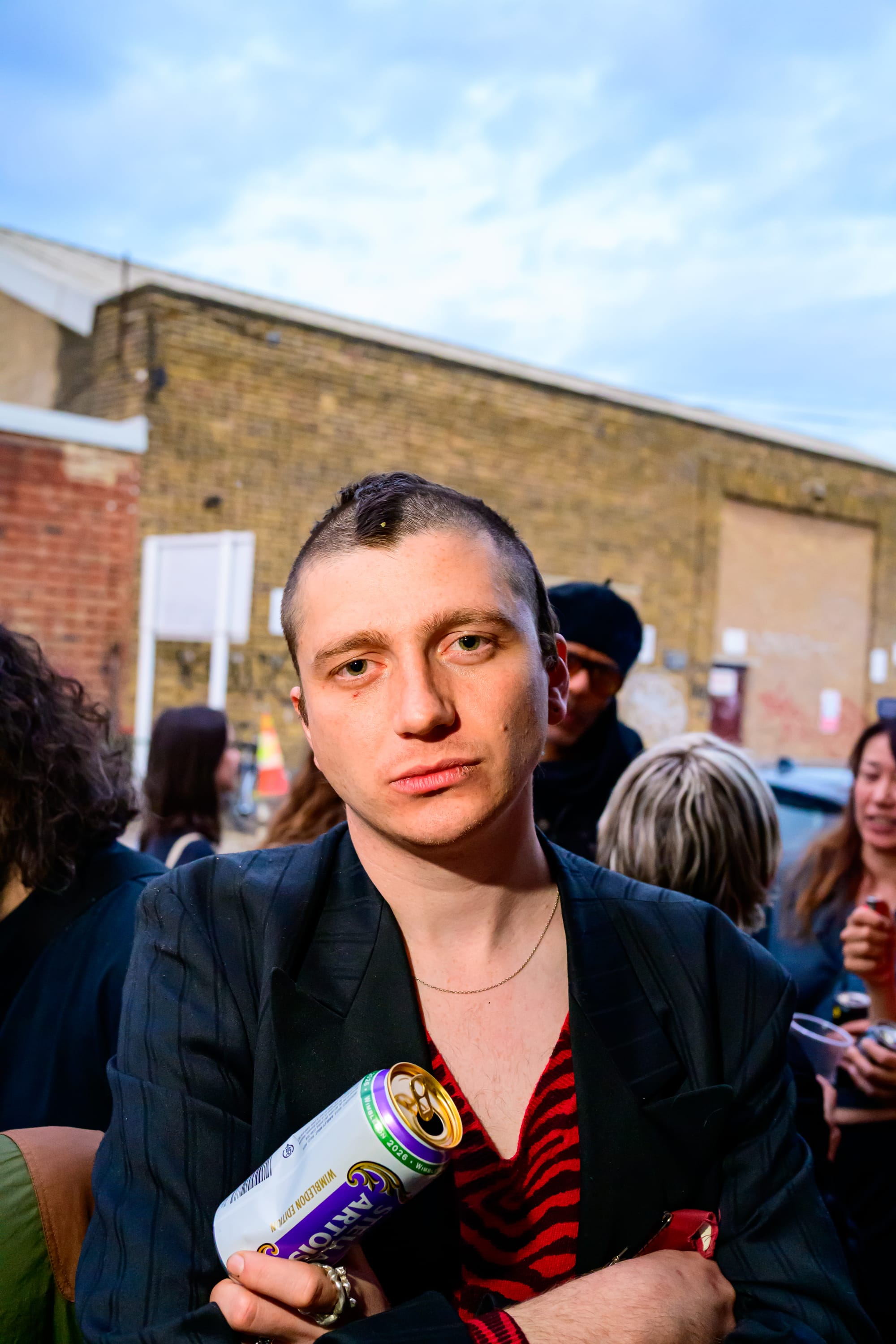A person with a strange haircut holding a can of Stella and crossing their arms.